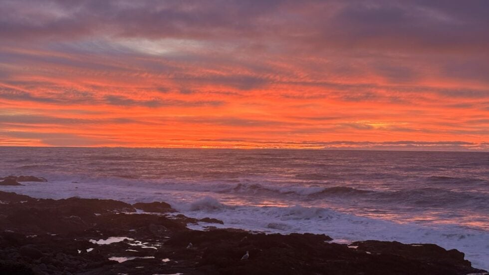 A bright pink and orange sunset contrasts with the dark rocky shoreline and white waves from the Adobe Resort in Yachats, Oregon.